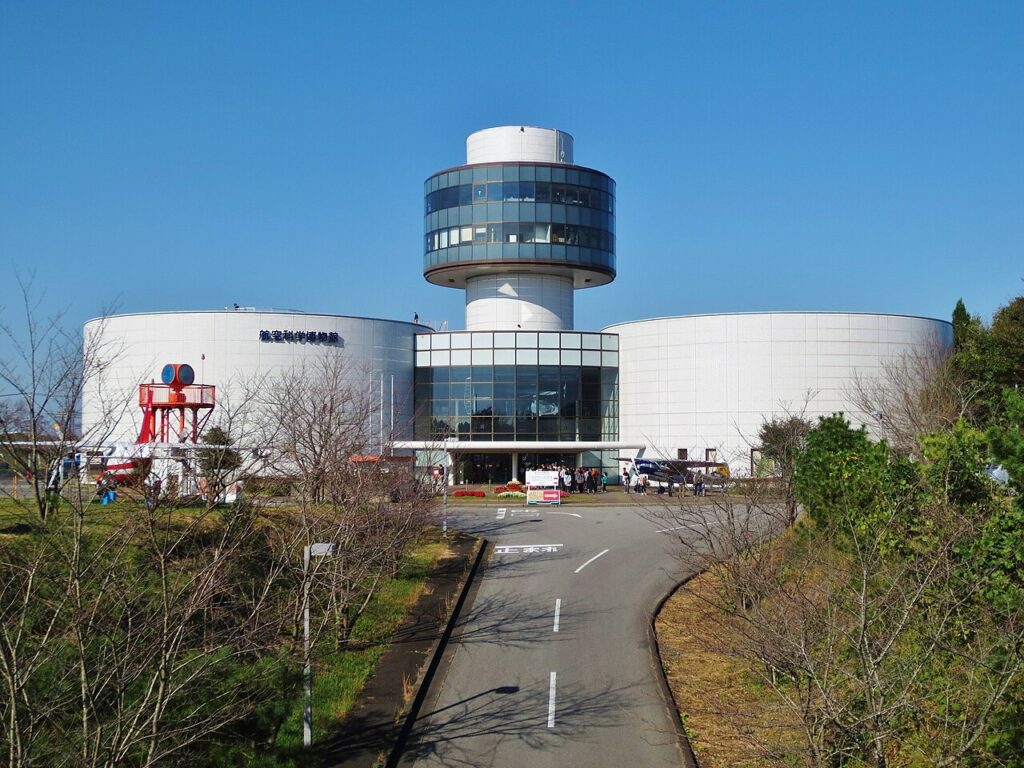 航空科学博物館の外観。白い円筒形の建物2棟の中央に、円形の窓が並ぶ展望塔が立つ。手前は駐車場から続く舗装路で、左手に赤いレーダーの展示、右手にヘリコプターが見える