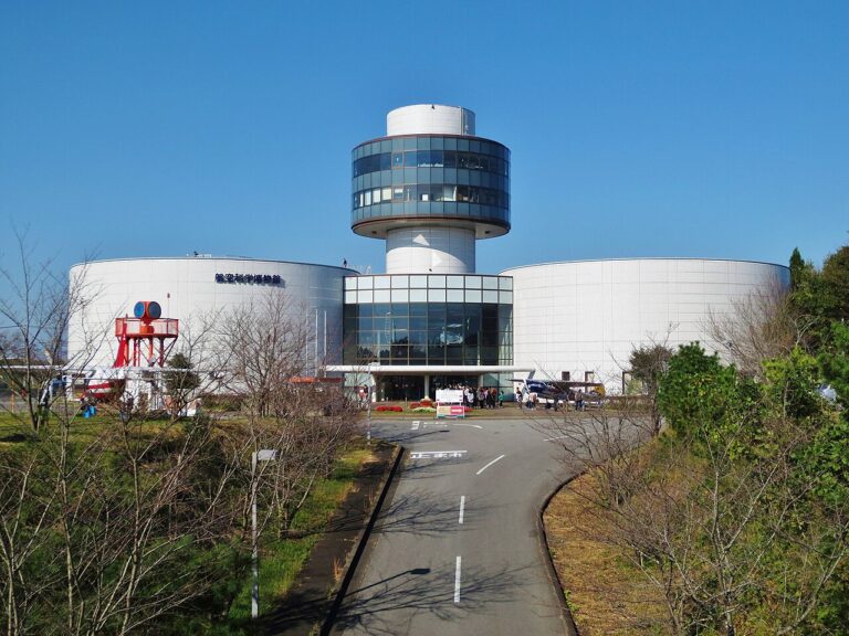 航空科学博物館の外観。白い円筒形の建物2棟の中央に、円形の窓が並ぶ展望塔が立つ。手前は駐車場から続く舗装路で、左手に赤いレーダーの展示、右手にヘリコプターが見える