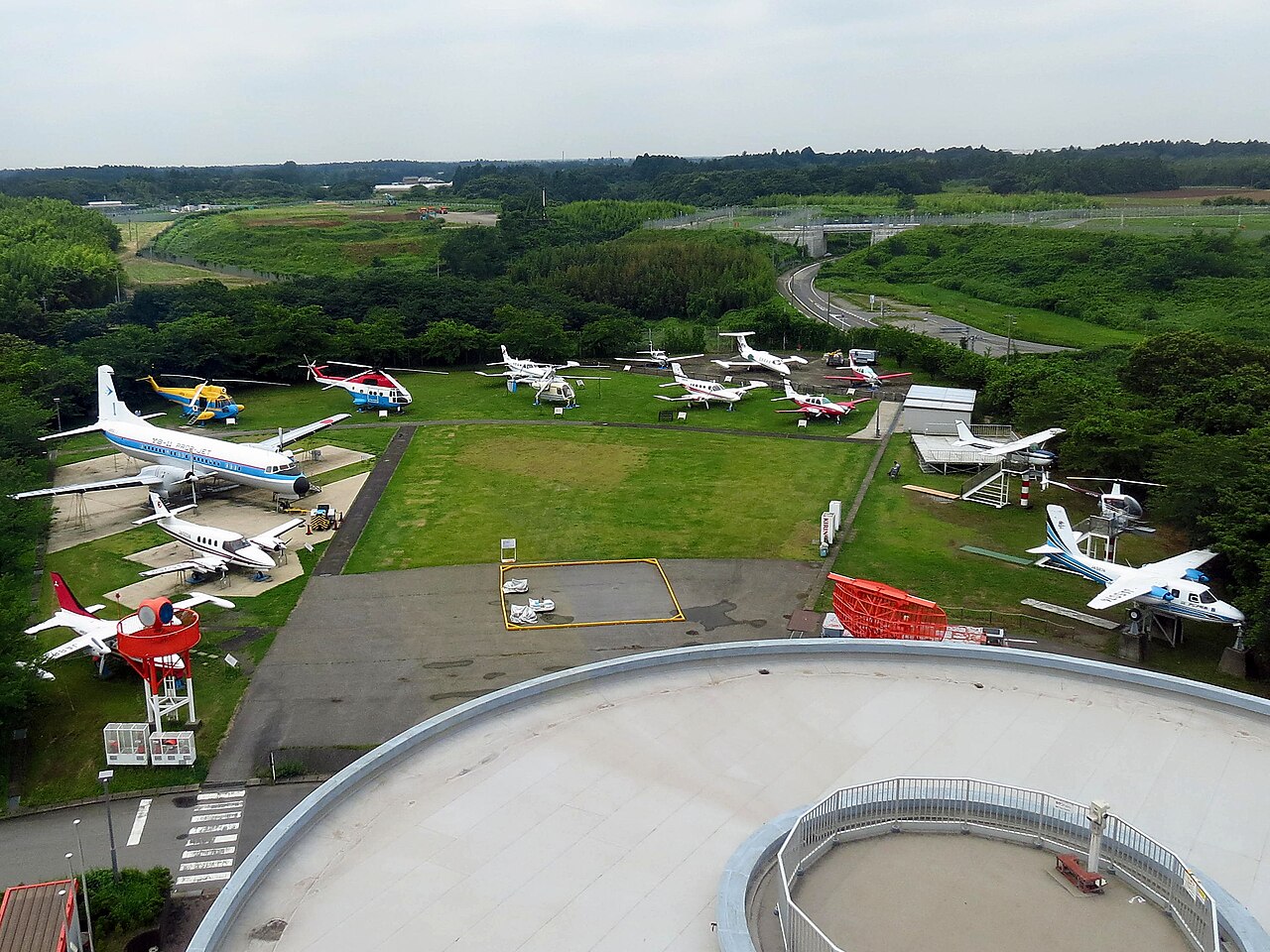 展望塔の上から見下ろした屋外展示場。芝生と舗装地の上に小型旅客機、双発機、ヘリコプター十数機が整然と並び、手前には博物館の白い円形の屋根と手すり付きの通路が写る。奥は林と道路