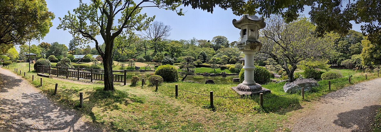日本庭園のパノラマ写真。中央に池と飛び石、右手前に大きな石灯籠が立ち、刈り込まれた低木と松が池を囲む。左手には木製の柵と橋、周囲は新緑の樹林に囲まれている