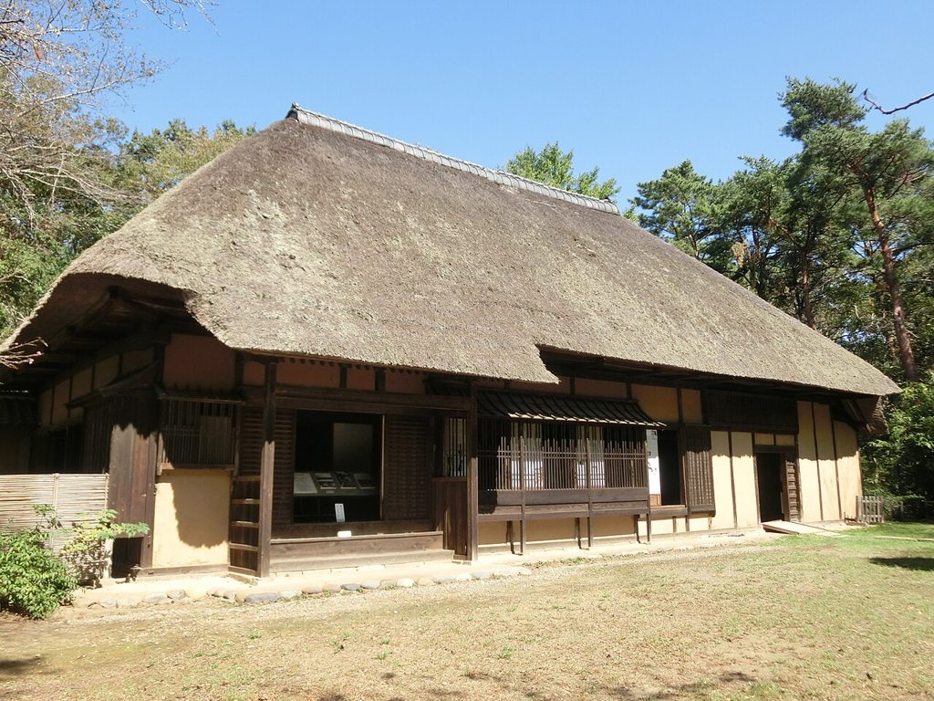 茅葺き屋根の大きな古民家。正面に濡れ縁と障子、格子戸が並び、軒下に展示解説のパネルが置かれている。建物の周囲は芝地で、背後に松などの高い木立が迫る