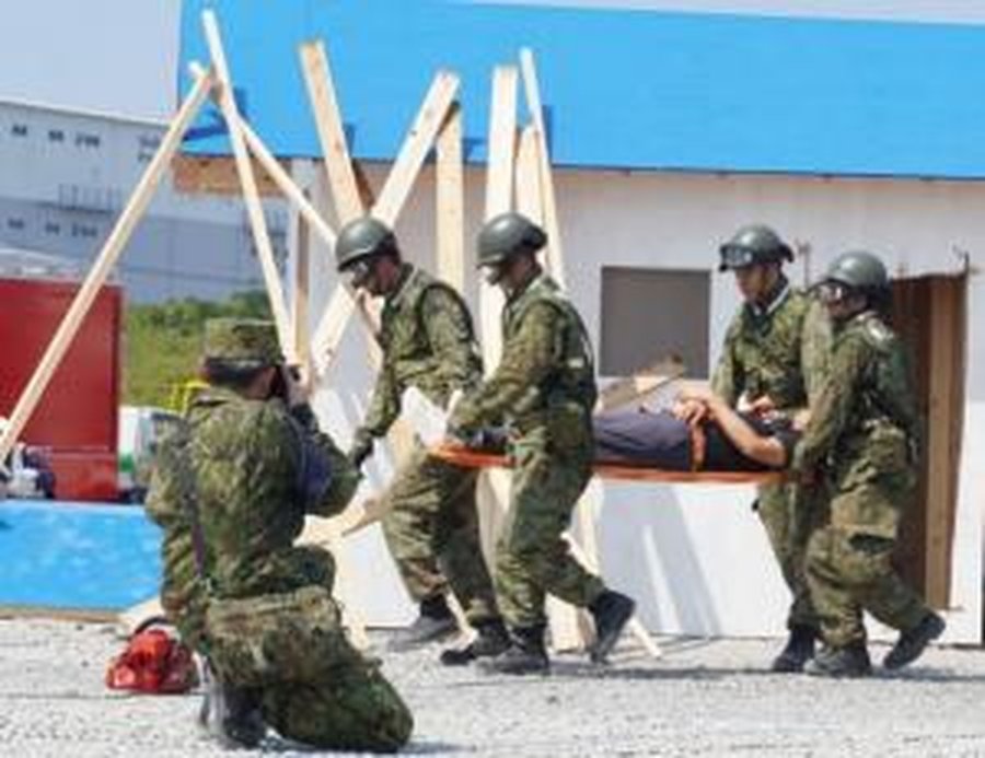 迷彩服にヘルメット姿の陸上自衛隊員5名が、負傷者役の人を乗せた担架を運んでいる。背景に倒壊家屋を模した角材とプレハブ状の構造物、青い仮設フェンスが見える