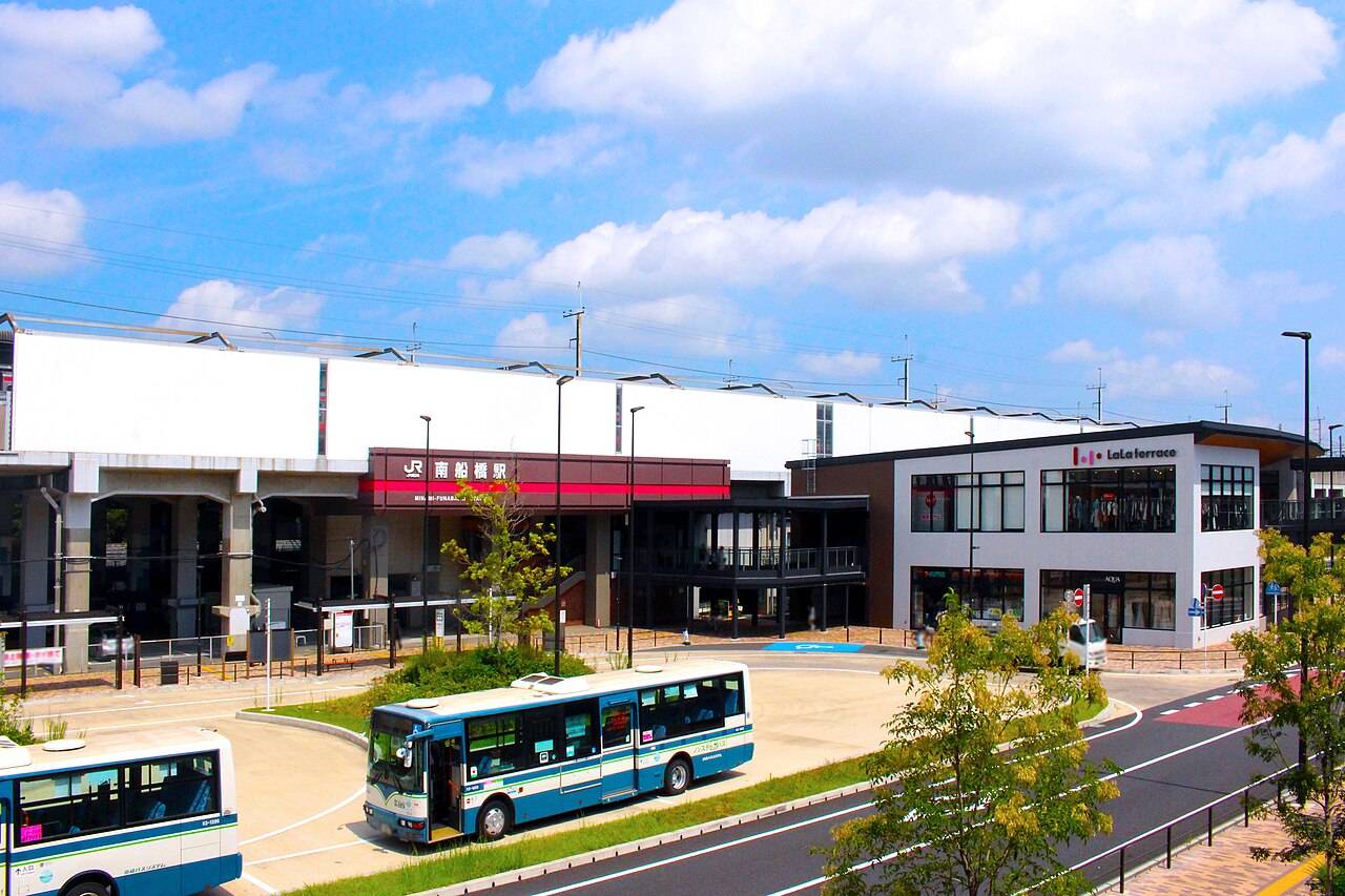 青空の下のJR南船橋駅を高い位置から見下ろした風景。中央に赤い帯に白抜きで「南船橋駅」と書かれた横長の駅舎、その背後に白い高架の線路が長く伸びる。右手に「LaLa terrace」の看板を掲げた2階建ての商業施設、手前のロータリーには青と白の路線バスが2台停まっている