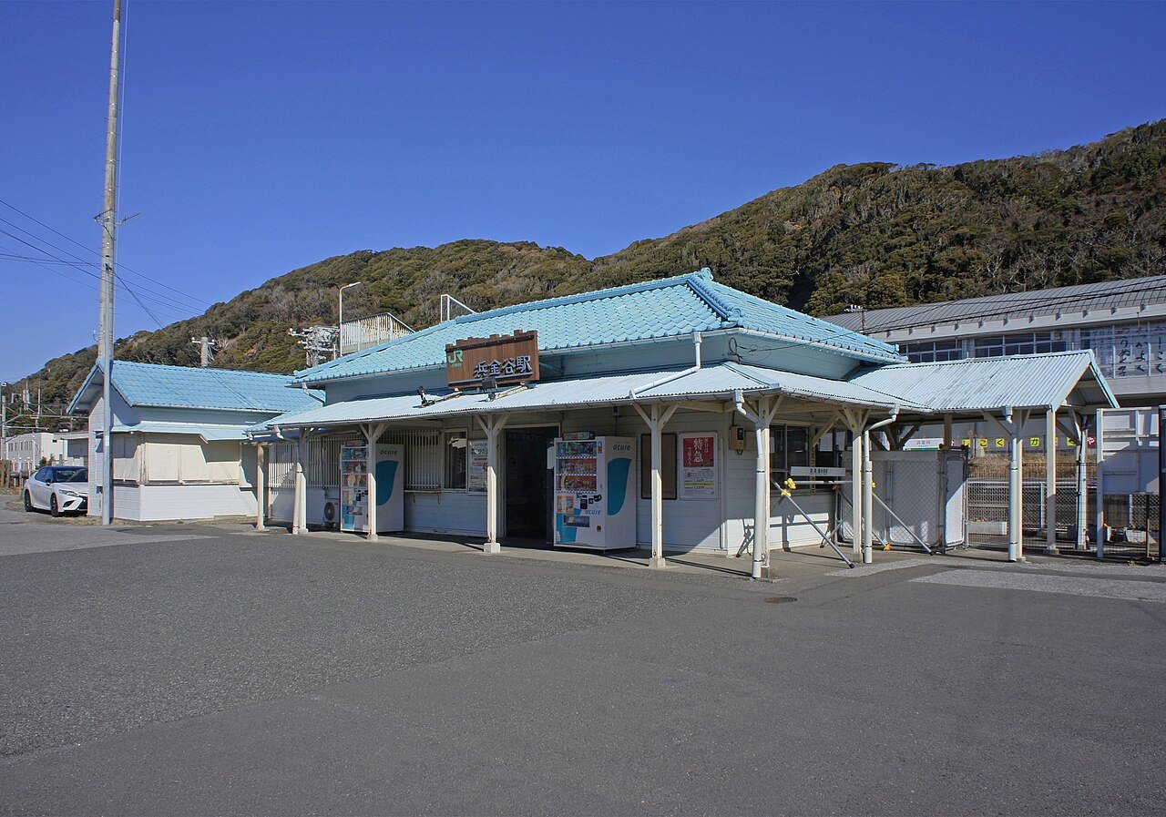 JR内房線・浜金谷駅の駅舎を正面から見た写真。水色の瓦屋根の小さな平屋で、軒下に木製の「浜金谷駅」看板と自動販売機が2台、右手に上屋とホームが続き、背後に低い山が迫る
