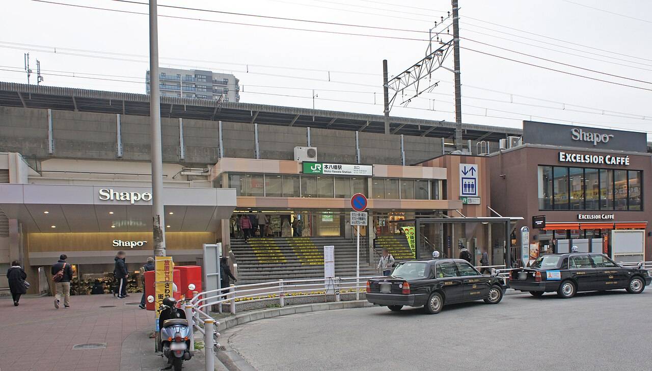 JR本八幡駅北口。高架下にある改札口の上に緑色のJRマークと「本八幡駅 北口」の駅名標、右手に「Shapo」「EXCELSIOR CAFFÈ」の看板が並ぶ商業施設、手前のロータリーに黒いタクシーが2台停まる