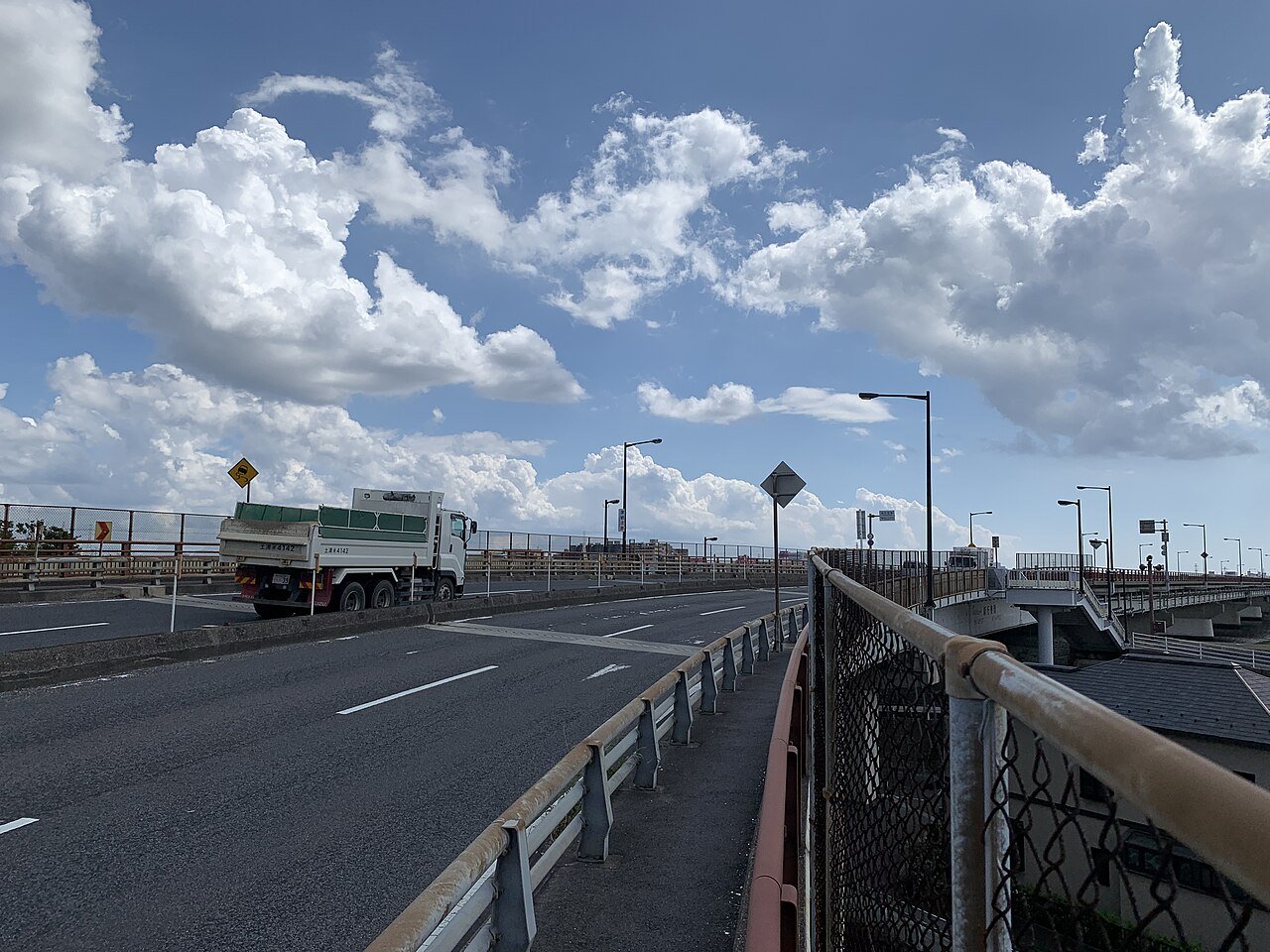 新行徳橋の上から見た風景。片側2車線の車道を緑の荷台のダンプカーが走り、右側にガードレールと金網フェンスに挟まれた歩道が奥へ続く。空には夏の入道雲が大きく広がる