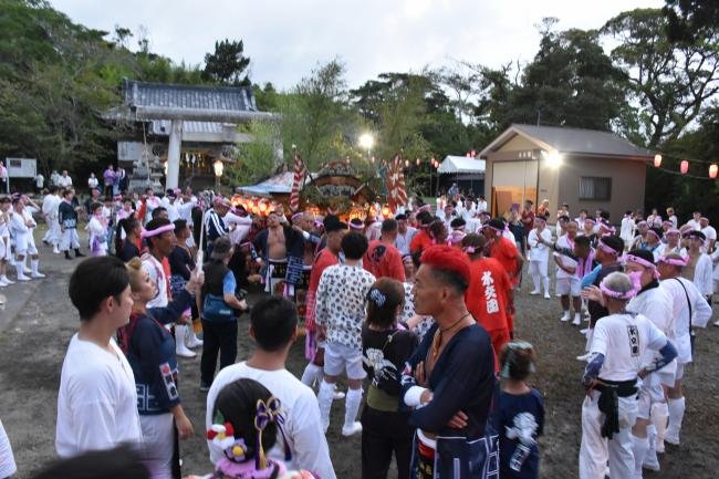 夕暮れの神社境内。石鳥居の手前に提灯を灯した屋台が据えられ、その周りを白装束や赤い法被の担ぎ手、浴衣姿の見物人が大勢取り囲んでいる。背景は木立と社務所の建物