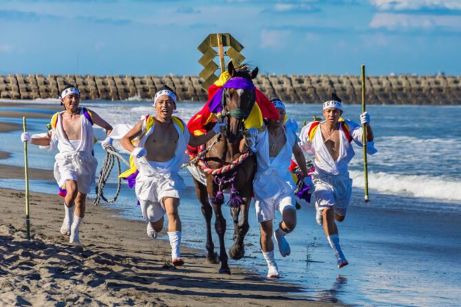 砂浜を黒毛の神馬が全力で疾走し、その前後を白装束に白鉢巻の男性5人が並走している。馬の背には金色の御幣が立てられ、赤と黄の錦の馬飾りがかけられている。背景は消波ブロックの堤防と白波