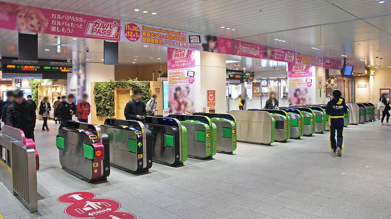 JR京葉線 海浜幕張駅の改札口。10通路以上の自動改札機が横一列に並び、天井から発車標が下がる。改札の上と柱にはピンク色のゲームイベントの横断幕と広告がびっしり掲げられ、右奥に売店。数人の乗客が改札に向かって歩いている