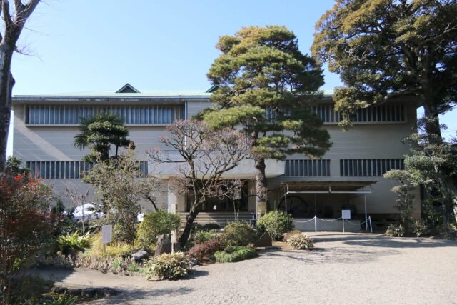野田市郷土博物館の外観。左右対称の白い横長の建物で、上部に正倉院の校倉造を思わせる水平材が並ぶ。屋根からは小さな三角屋根が突き出し、手前は砂利敷きの前庭に松と落葉した木が立つ