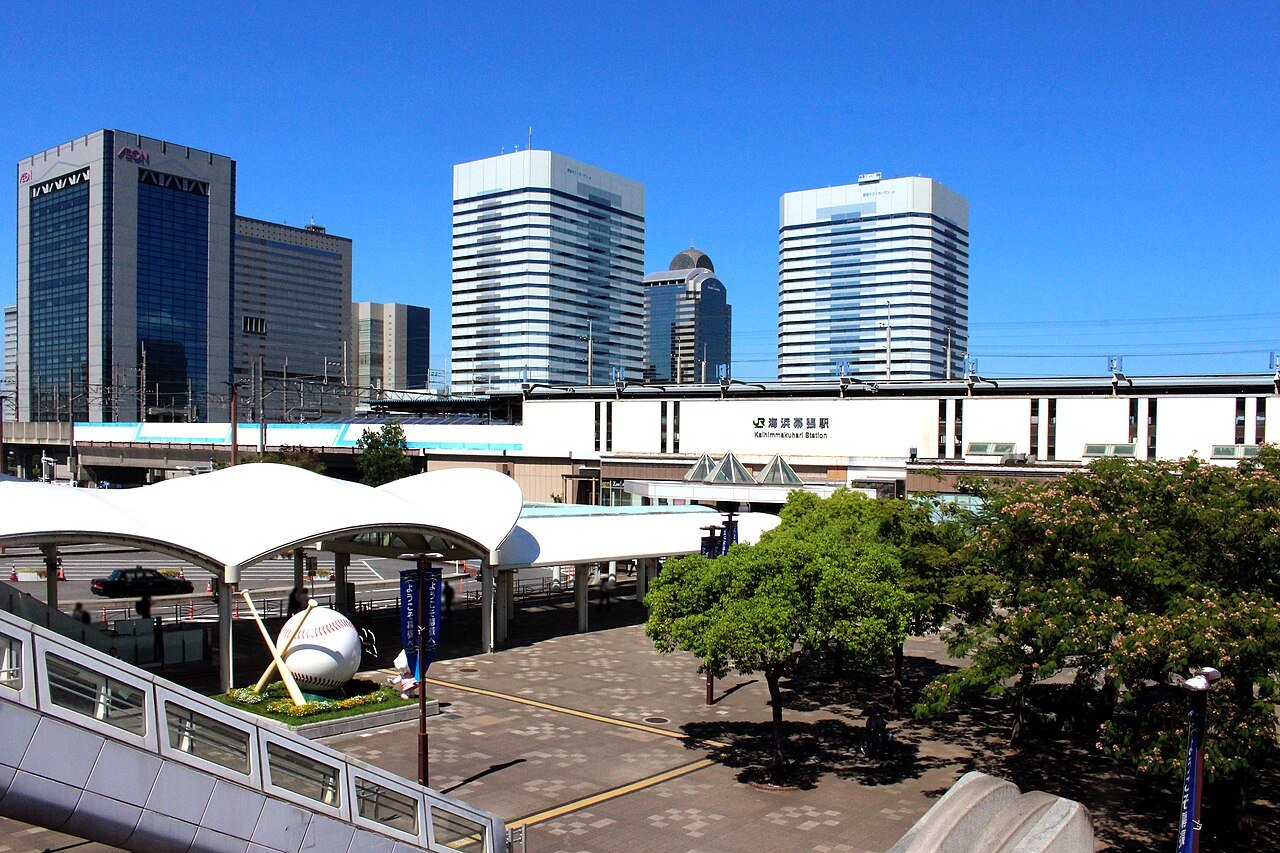 青空の下の海浜幕張駅を高い位置から見た風景。中央に「海浜幕張駅 Kaihimmakuhari Station」と書かれた白い駅舎が横に伸び、その後ろにイオンや幕張テクノガーデンの高層ビルが並ぶ。手前の駅前広場には白い膜屋根のバスロータリー、バットとボールをかたどった野球のモニュメント、街路樹