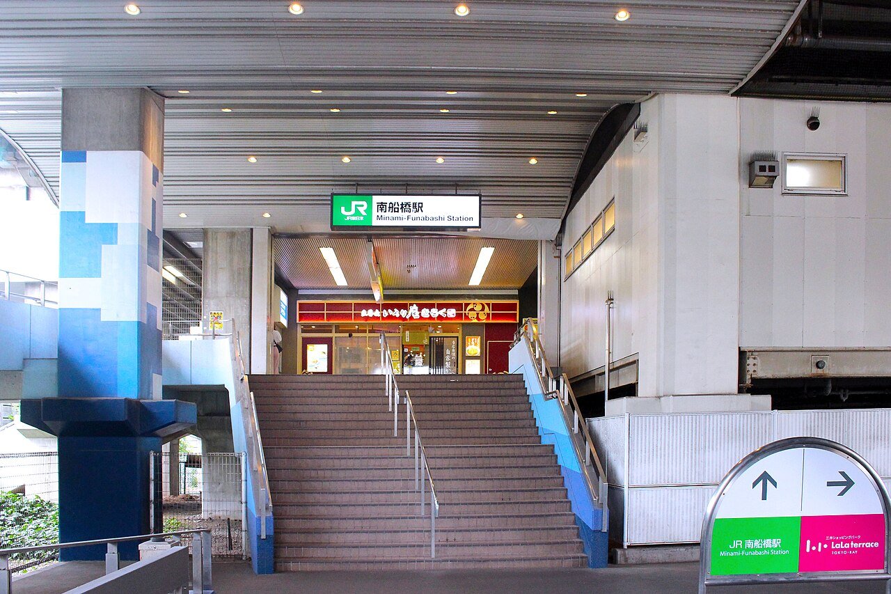 JR南船橋駅の出入口。高架下の天井の低い通路の奥に幅の広いコンクリートの階段が上へ伸び、その上に「JR 南船橋駅 Minami-Funabashi Station」の緑の駅名標が掲げられている。階段の上には飲食店の赤い看板。右手前に「JR南船橋駅」と「LaLa terrace TOKYO-BAY」の方向を示す矢印看板が立つ