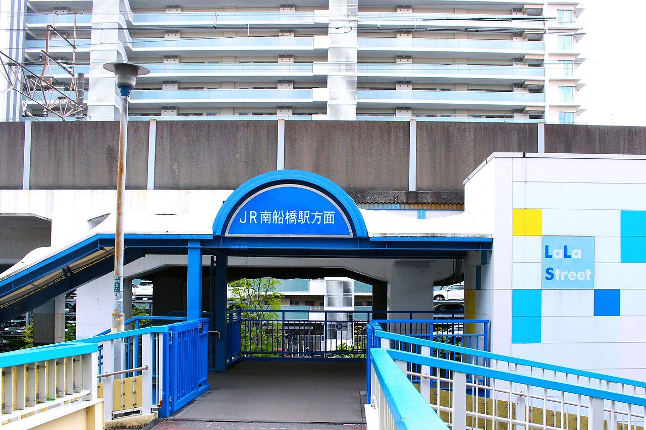 「JR南船橋駅方面」と書かれた青いアーチ状の看板が掲げられた高架下の通路の入口。左に青い手すりの階段、右の壁面に「LaLa Street」のロゴと水色・黄色のタイル。奥にコンクリートの高架橋と高層マンションが見える