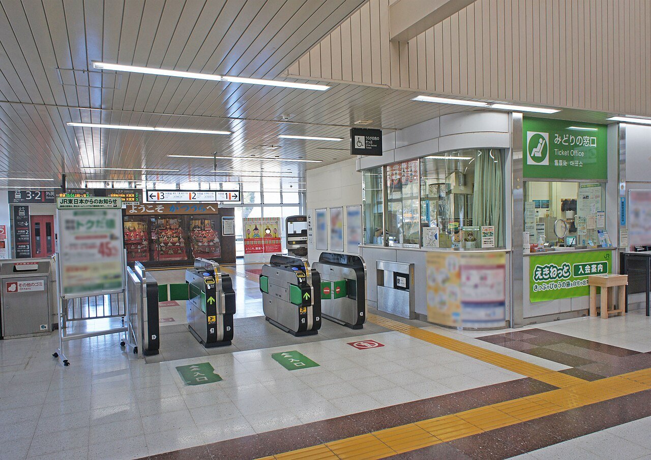 JR勝浦駅の改札口。中央に3通路の自動改札機が並び、右手に緑の看板の「みどりの窓口」、左の壁際には「ようこそ かつうらへ」の看板の下にひな人形を並べたガラスケースが置かれている。天井から外房線の発車標が下がり、人はいない