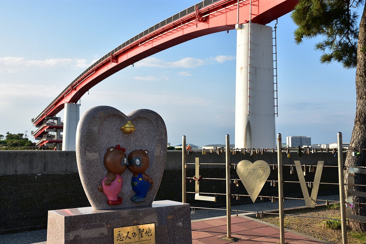 青空を背景に、赤い中の島大橋が大きく弧を描いて伸びる。手前に「恋人の聖地」と刻まれたハート型の御影石のモニュメントがあり、ピンクとブルーの服を着た狸のカップル像が向き合う。右側の柵には南京錠がびっしり掛かったLOVEのオブジェ