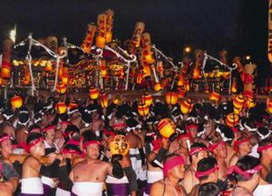 夜。提灯を無数に灯した神輿が何基も横一列に並び、オレンジ色に光っている。手前にはピンクや赤の鉢巻きを締めた担ぎ手が大勢ひしめいている