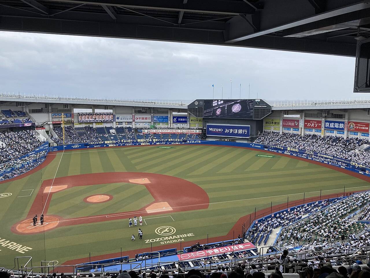 スタンド上段の屋根の下からグラウンドを見下ろした眺め。天然色の内野の土と縞模様の外野の芝、中央奥に大型ビジョンとスコアボード、左右のスタンドには観客が入っている。画面上部には屋根の黒い鉄骨の梁が横切り、その先に曇り空が見える