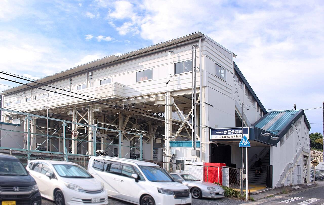 京成本線・宗吾参道駅の外観。白い壁の高架駅舎が横に伸び、その下に線路を支える鉄骨の架構が見える。右手の入口の上に「京成電鉄 宗吾参道駅 Sōgosandō Station」と書かれた紺色の駅名看板が掲げられ、その下から階段が上へ続く。手前の駐車スペースには数台の乗用車が停まり、道路は右手前へ下っている