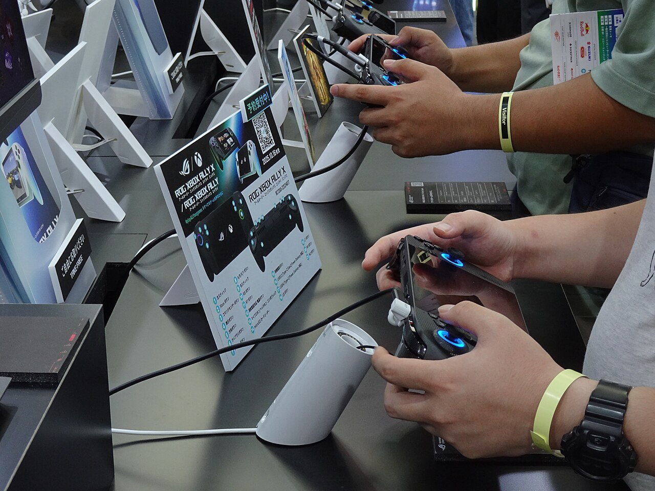 展示ブースの試遊卓で、来場者2人が携帯型ゲーム機のコントローラーを両手で握っている。手首には黄色い「Visitor」のリストバンド。卓上に「予約受付中」のQRコード付きPOPと、ROG Xbox Ally X の操作説明ボードが立てられている