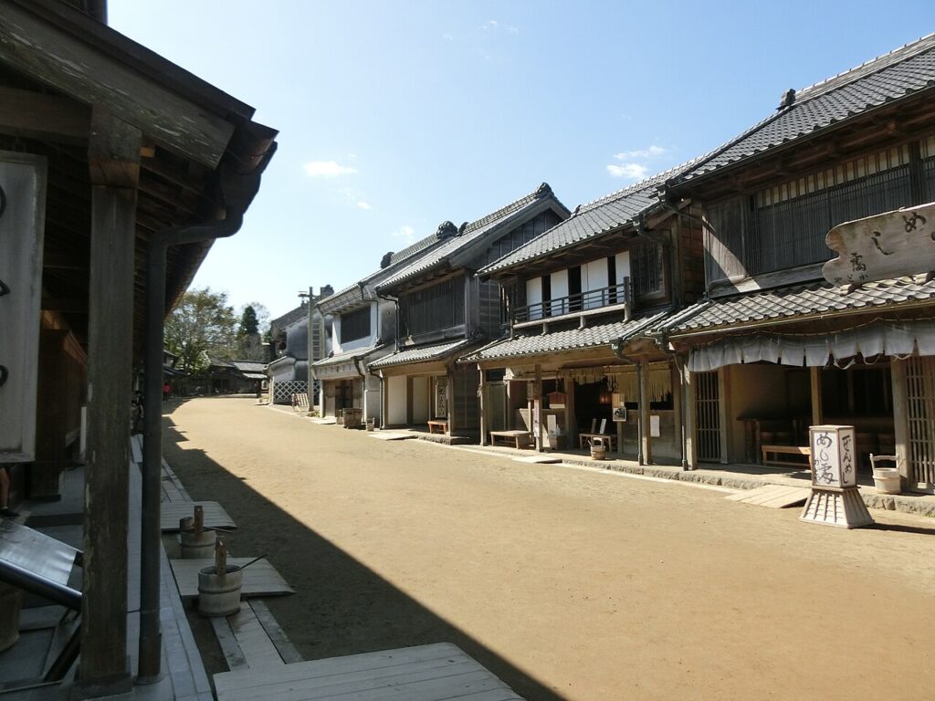 房総のむらの商家の町並み。土の道の両側に瓦屋根と蔵造りの町家が並び、右手の店先に「めし屋」と書かれた白い看板と暖簾が下がる。左手前は建物の軒と桶が置かれた土間