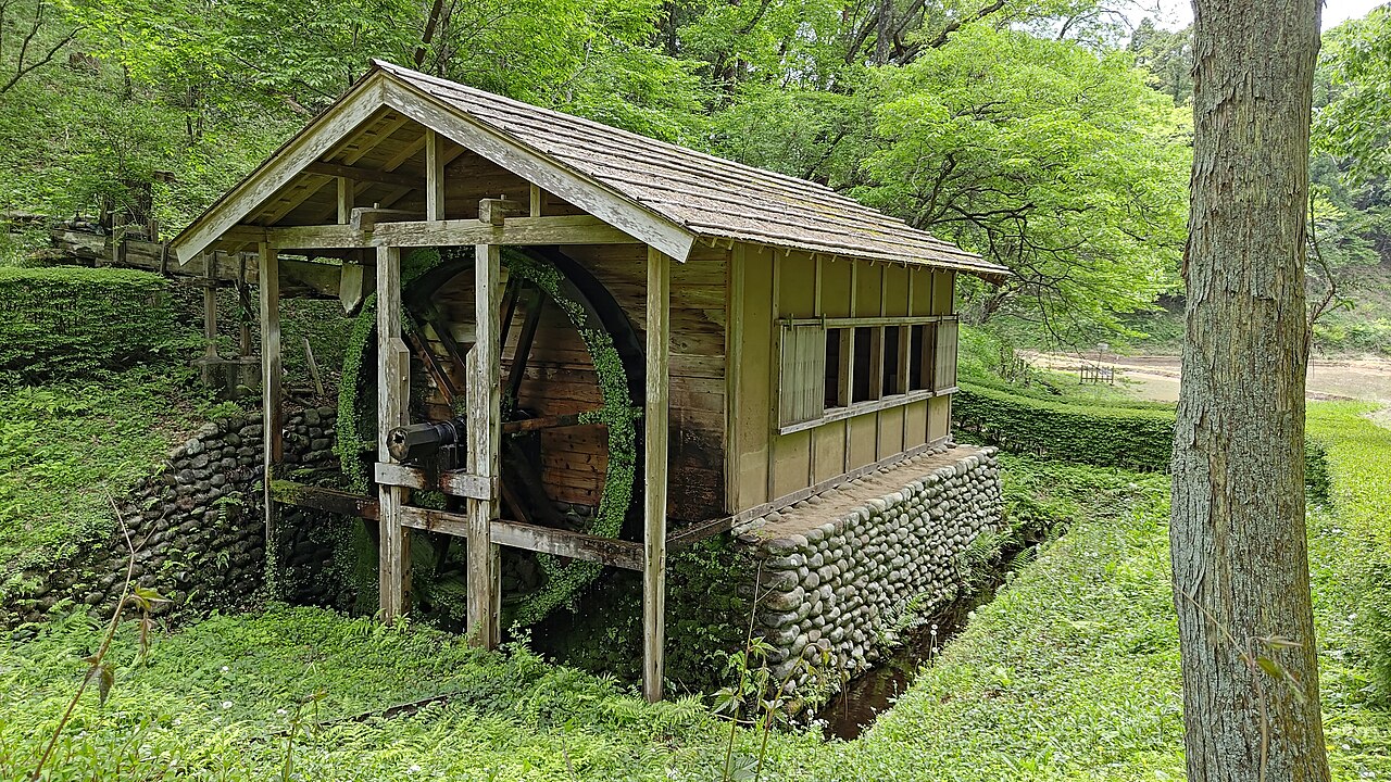 木立に囲まれた水車小屋。板葺きの屋根の下に大きな木製の水車が据えられ、石積みの土台の脇を細い水路が流れる。周囲は下草に覆われ、手前に太い樹木の幹が立つ