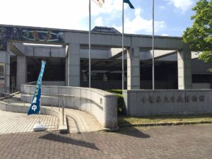 千葉県立中央博物館の正面入口。石張りの外壁に館名が刻まれ、青いのぼり旗と掲揚ポールが並ぶ