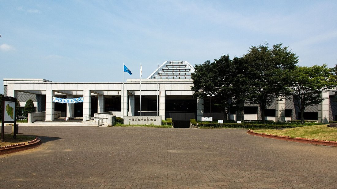 千葉県立中央博物館の全景。低層の白い建物が横に長く伸び、中央に三角形の屋根、手前に広いレンガ敷きの前庭が広がる
