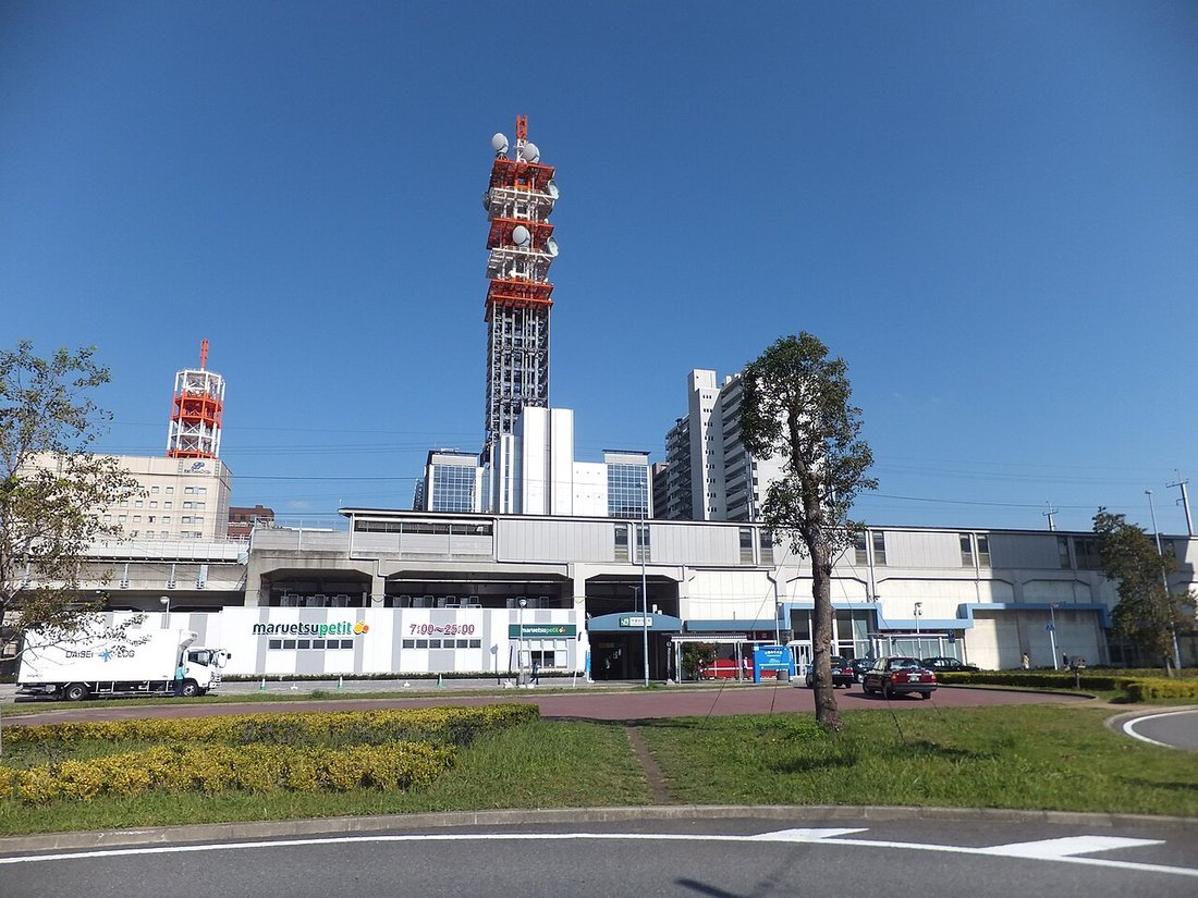 千葉みなと駅の駅前。高架の駅舎とスーパーの看板、背後に赤白の電波塔とマンション、手前はロータリーの植え込みと車道