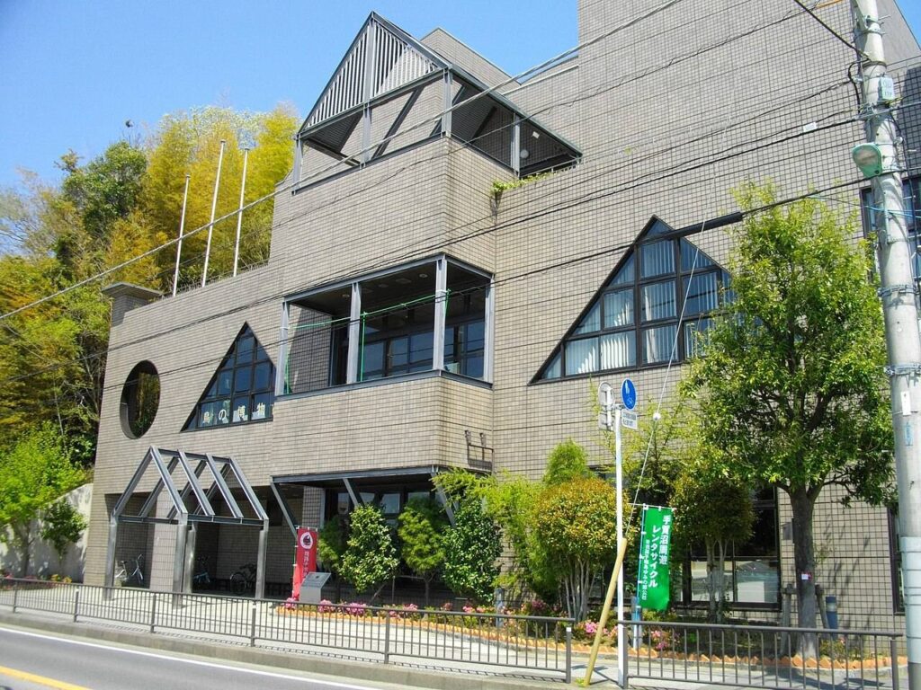 我孫子市鳥の博物館の外観。ベージュのタイル張りの3階建てで、大小の三角形をかたどった窓や屋上構造物、壁面の丸窓が並ぶ。道路側の壁に「鳥の博物館」の文字があり、手前の歩道には手賀沼周遊レンタサイクルの緑ののぼりが立つ