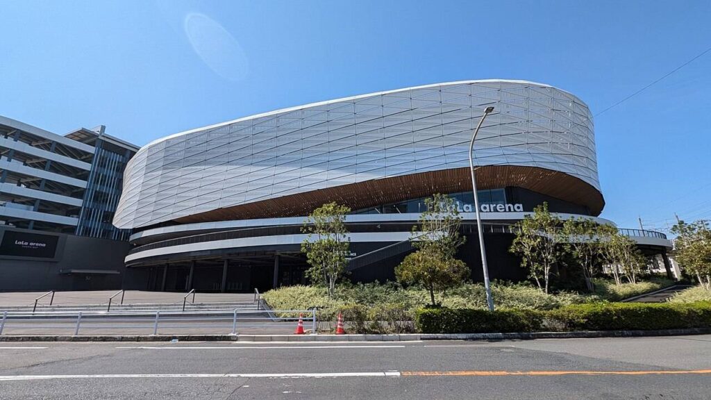 青空の下のLaLa arena TOKYO-BAYの外観。楕円形に膨らんだ銀色のメッシュ状の外壁が大きく張り出し、その下端に木目調の庇と黒いガラス面が回り込む。「LaLa arena」の白い立体文字が壁面に掲げられ、手前に街路樹と歩道、右奥に立体駐車場と歩道橋が見える