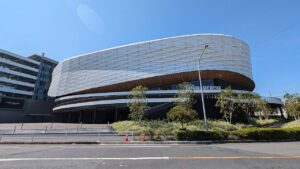 青空の下のLaLa arena TOKYO-BAYの外観。楕円形に膨らんだ銀色のメッシュ状の外壁が大きく張り出し、その下端に木目調の庇と黒いガラス面が回り込む。「LaLa arena」の白い立体文字が壁面に掲げられ、手前に街路樹と歩道、右奥に立体駐車場と歩道橋が見える