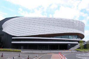 曇りがちな空の下のLaLa arena TOKYO-BAYの外観を正面から捉えた写真。白いメッシュ状のパネルで覆われた楕円形の巨大な屋根が横に大きく張り出し、その下に木目調の庇と2層の白い回廊が回る。右端の壁面に「LaLa arena」の白い文字と「2F STAND ENTRANCE」の表示。手前は舗装された広場と赤白のポール