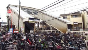 京成菅野駅の出入口。白い三角屋根の駅舎から長い跨線橋が右へ伸び、庇の下に「京成電鉄 菅野駅 Sugano Station」の駅名看板が掛かる。手前は歩道いっぱいに停められた自転車の群れで、左に赤い消火栓の標識が立つ