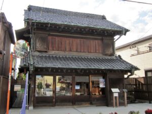 木造二階建ての商家。瓦屋根が二段に重なり、二階の正面は太い木の板戸で覆われ、深い軒が張り出す。一階は間口いっぱいのガラス戸で、内部に神輿の飾り金具や道具が見える。右手前に木の解説板が二枚立ち、前面は石畳