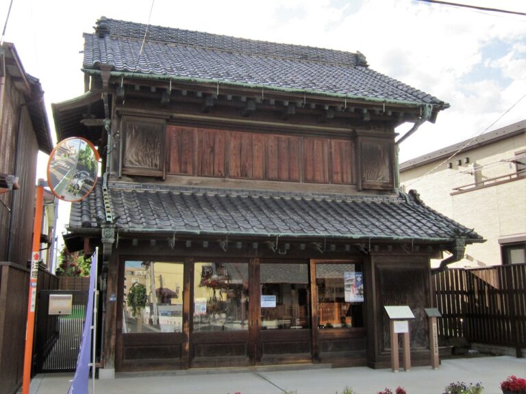 木造二階建ての商家。瓦屋根が二段に重なり、二階の正面は太い木の板戸で覆われ、深い軒が張り出す。一階は間口いっぱいのガラス戸で、内部に神輿の飾り金具や道具が見える。右手前に木の解説板が二枚立ち、前面は石畳
