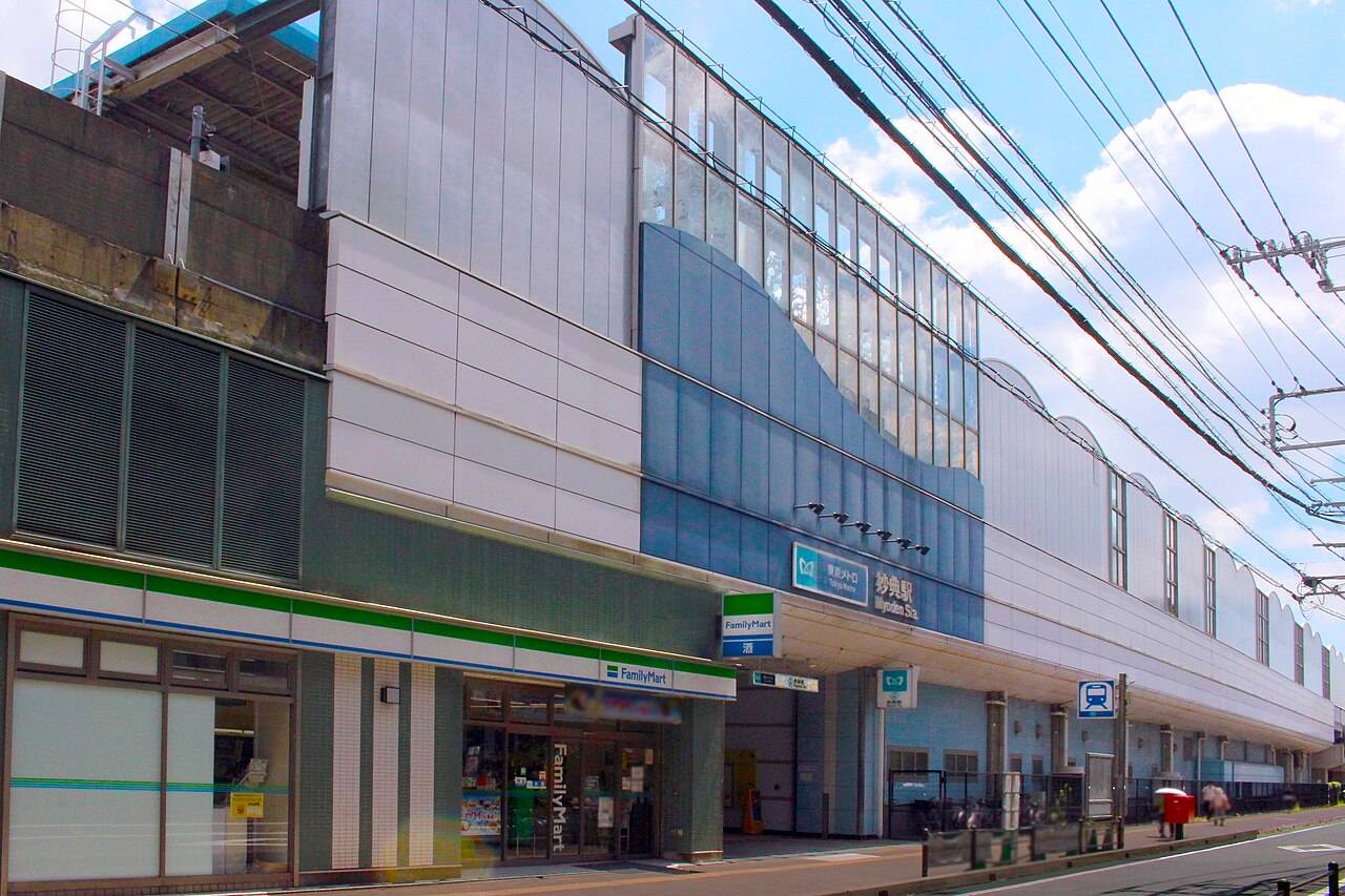 東京メトロ東西線・妙典駅の高架駅舎を北側から見上げた写真。銀と青の波形パネルで覆われた壁面に「東京メトロ 妙典駅 Myoden Sta.」の駅名サインが付き、1階にはファミリーマートが入る。上空に架線と夏の青空