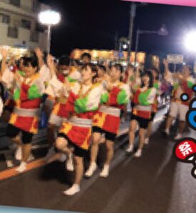 夜の県道で赤と緑の法被を着た踊り手が列になって上総おどりを踊っている。街灯と提灯が並ぶ通りに大勢が並ぶ