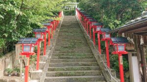 遠見岬神社の石段。苔むした灰色の石段がまっすぐ奥へ伸び、左右に朱塗りの木製灯篭が10基以上ずらりと並ぶ。灯篭の柱には奉納者名の木札が下がる。左手前に「富咲の段」と書かれた木の看板があり、石段の上には注連縄が渡され、周囲は濃い緑の木々に覆われている