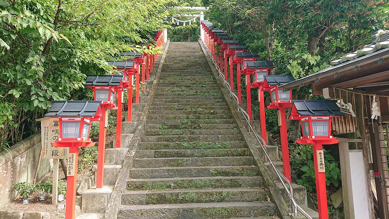 遠見岬神社の石段。苔むした灰色の石段がまっすぐ奥へ伸び、左右に朱塗りの木製灯篭が10基以上ずらりと並ぶ。灯篭の柱には奉納者名の木札が下がる。左手前に「富咲の段」と書かれた木の看板があり、石段の上には注連縄が渡され、周囲は濃い緑の木々に覆われている