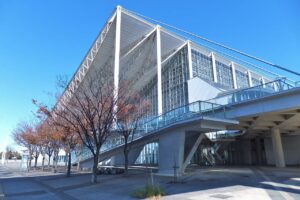青空の下の幕張メッセ国際展示場9〜11ホールの外観。白い鉄骨の支柱が並び、大きく張り出した三角形の白い屋根とガラス張りの壁面が斜めに伸びる。手前に葉を落としかけた並木と歩道、右手に高架のペデストリアンデッキ