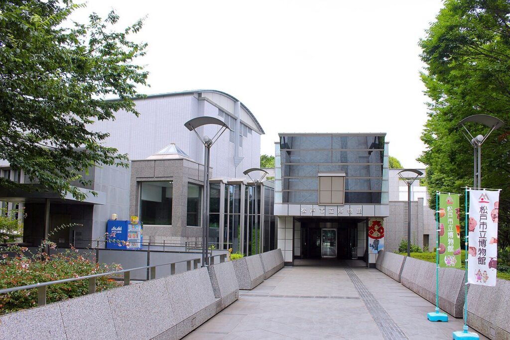 松戸市立博物館の正面エントランス。ガラス張りの高い庇の下に「松戸市立博物館」の看板と自動ドアがあり、白い外壁とかまぼこ型の屋根が奥に続く。石畳のアプローチの右手に、館のキャラクターが描かれた緑色ののぼりが2本立っている