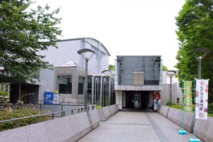 松戸市立博物館の正面エントランス。ガラス張りの高い庇の下に「松戸市立博物館」の看板と自動ドアがあり、白い外壁とかまぼこ型の屋根が奥に続く。石畳のアプローチの右手に、館のキャラクターが描かれた緑色ののぼりが2本立っている