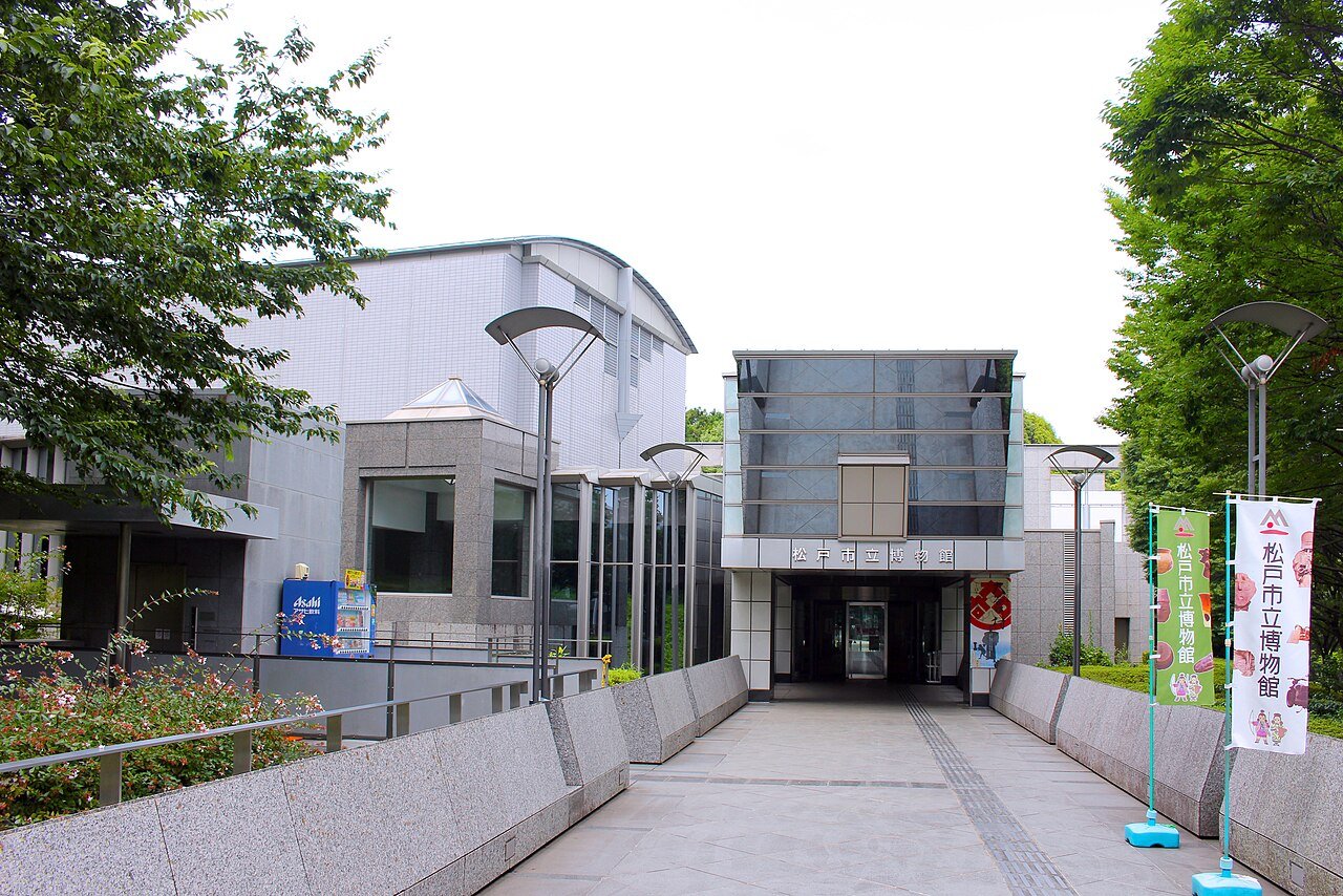 松戸市立博物館の正面エントランス。ガラス張りの高い庇の下に「松戸市立博物館」の看板と自動ドアがあり、白い外壁とかまぼこ型の屋根が奥に続く。石畳のアプローチの右手に、館のキャラクターが描かれた緑色ののぼりが2本立っている