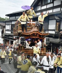成田山表参道を進む木造の山車。屋根の上に法被姿の男性3人が立ち、一人が紫の日傘を差している。山車の左右には「田」の一文字を大書した白い提灯が吊るされ、彫刻の施された欄間の下に囃子方が座る。黄色い法被の曳き手が大勢で綱を引き、背景は表参道の瓦屋根の町家