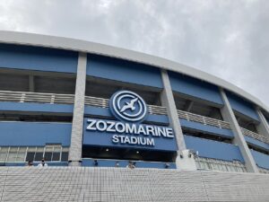 見上げる角度で撮ったZOZOマリンスタジアムの外壁。青と水色の横縞のスタンド壁面に、カモメをかたどった円形エンブレムと白い立体文字の「ZOZOMARINE STADIUM」が掲げられている。曇り空。手前はタイル張りの外周デッキ