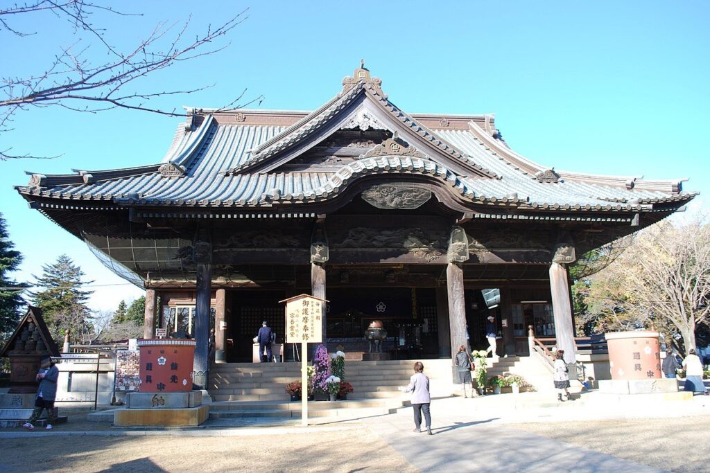 青空の下に建つ宗吾霊堂の大本堂。入母屋屋根を重ねた大きな木造建築で、灰色の瓦屋根と、軒下に龍などの彫刻を施した欅の梁が見える。正面に石段と賽銭箱、中央に金属製の香炉が置かれ、手前に「御護摩奉修 宗吾霊堂」と書かれた木の立て札。石段の脇に菊の鉢が並び、参拝者が数人歩いている