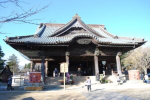 青空の下に建つ宗吾霊堂の大本堂。入母屋屋根を重ねた大きな木造建築で、灰色の瓦屋根と、軒下に龍などの彫刻を施した欅の梁が見える。正面に石段と賽銭箱、中央に金属製の香炉が置かれ、手前に「御護摩奉修 宗吾霊堂」と書かれた木の立て札。石段の脇に菊の鉢が並び、参拝者が数人歩いている
