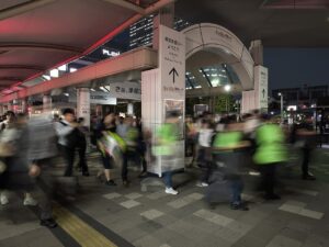 夜の海浜幕張駅前のペデストリアンデッキ。大勢の来場者が幕張メッセ方面へ向かって足早に歩き、動きがぶれて写っている。中央に「幕張新都心へようこそ」「幕張メッセ 500m」の白い案内柱、奥に「さあ、準備は」と書かれた東京ゲームショウの横断幕。天井の膜屋根が赤く照らされている