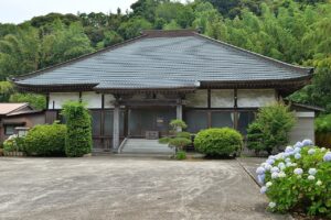 広済寺の本堂を正面から見た写真。緩やかに反った大きな瓦屋根の下に木の柱と白壁が並び、中央の階段の先に本堂の入口がある。手前は広く開けた土の広場、右手前に青紫の紫陽花、背後の斜面は一面の竹林