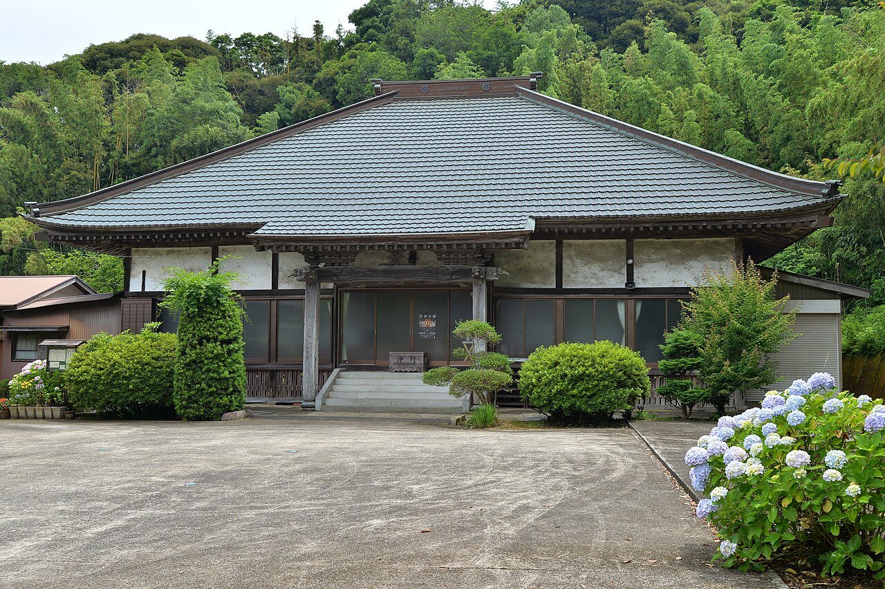 広済寺の本堂を正面から見た写真。緩やかに反った大きな瓦屋根の下に木の柱と白壁が並び、中央の階段の先に本堂の入口がある。手前は広く開けた土の広場、右手前に青紫の紫陽花、背後の斜面は一面の竹林